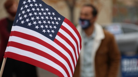 A handheld American flag is held in front of the camera. Slightly blurred in the background, we can see a man in a brown jacket with a white collar who is wearing a mask over his nose and mouth.