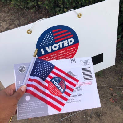 Someone holds an American flag with an "I voted" sticker on it, and an envelope for an absentee ballot